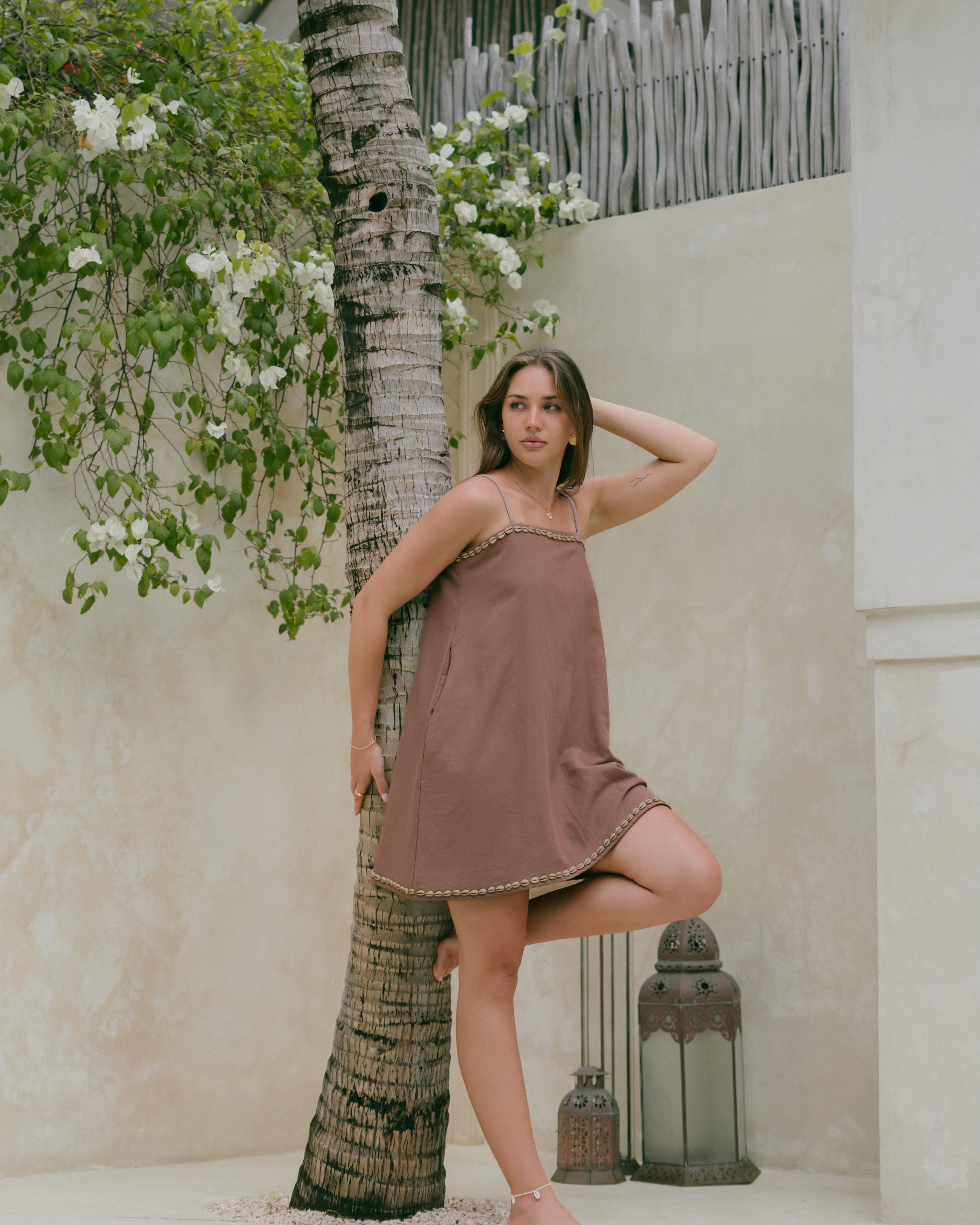 The cowrie shell mini dress features a relaxed shift silhouette, adjustable straps, and pockets — perfect for sun-filled days and coastal escapes.