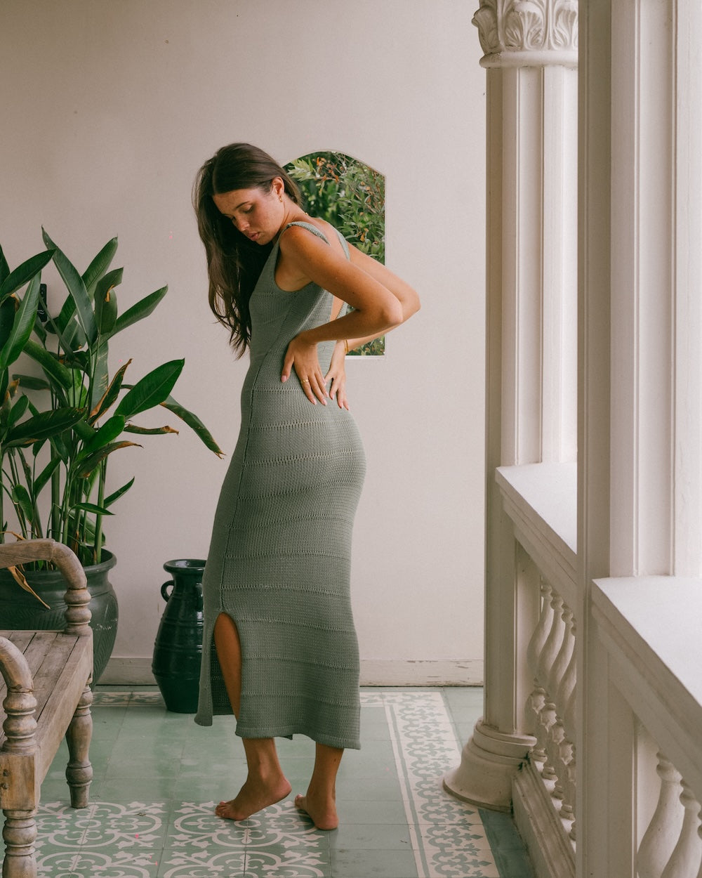 Noa Knit Dress in Sage, ribbed knit maxi dress with side slit, styled with one hand on hip against a plant-filled balcony