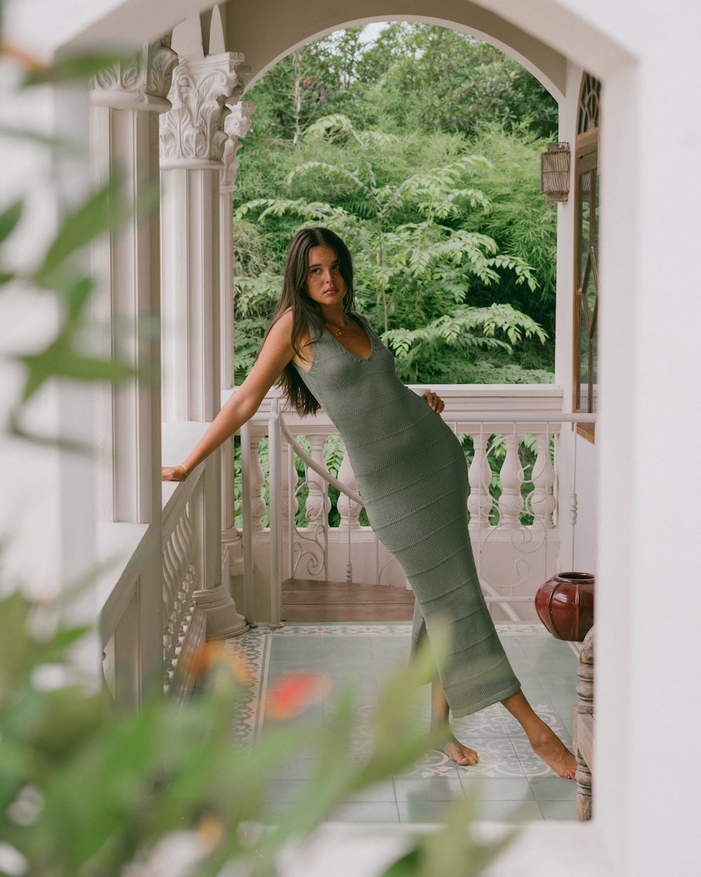 A woman wearing a sleeveless olive green ribbed midi dress stands barefoot on a white balcony with a lush green garden background, framed by an arched doorway.