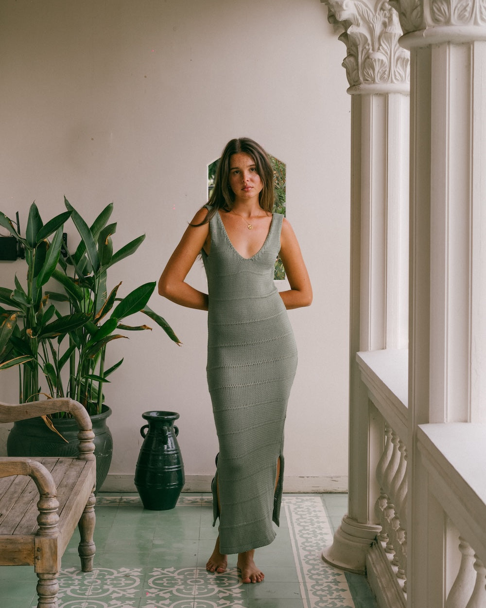 Noa Knit Dress in Sage, ribbed knit maxi dress with side slits, styled with one hand on hip against a plant-filled balcony