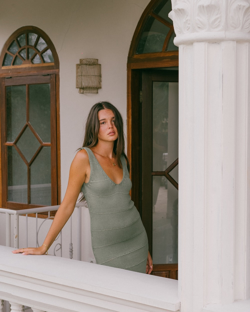 Noa Knit Dress in Sage, ribbed knit maxi dress with side slit, styled with one hand on hip against a plant-filled balcony