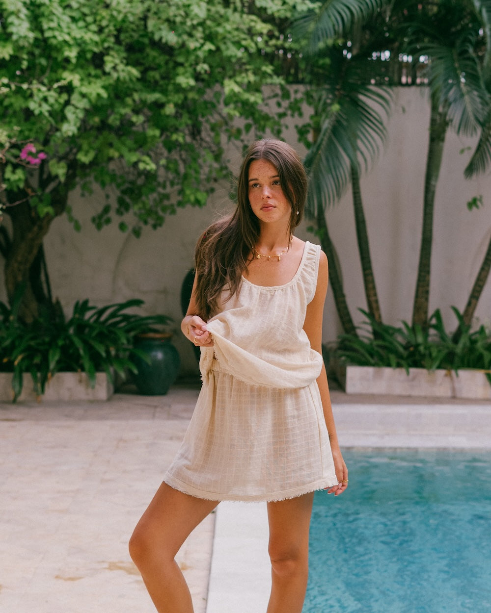 Sofia Singlet Top and Mini Skirt matching set in natural check ramie — model standing poolside holding skirt fabric, full-length front view showing gathered neckline and frayed hem — breathable natural fibre summer co-ord set — Malia The Label La Dolce Vita collection
