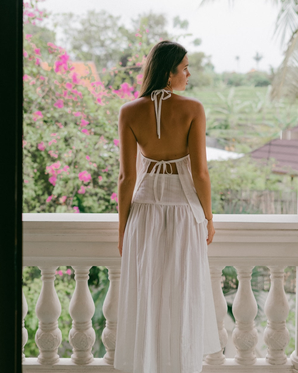 Sole Mare Singlet Top and Maxi Skirt rear view on balcony — full-length back view showing open back halter tie detail and floor-sweeping maxi skirt in white handloomed cotton, bougainvillea and tropical garden backdrop — backless halter top and maxi skirt set — Malia The Label
