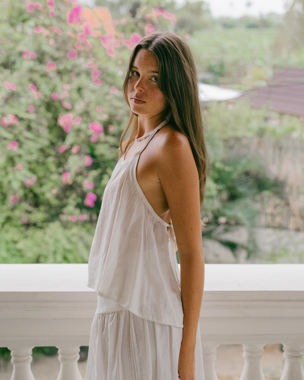 Sole Mare Singlet Top side portrait on balcony — three-quarter side view showing gathered halter neckline, open back silhouette and intricate handloomed cotton fabric, model looking over shoulder with floral backdrop — white cotton halter singlet top — Malia The Label slow fashion Australia