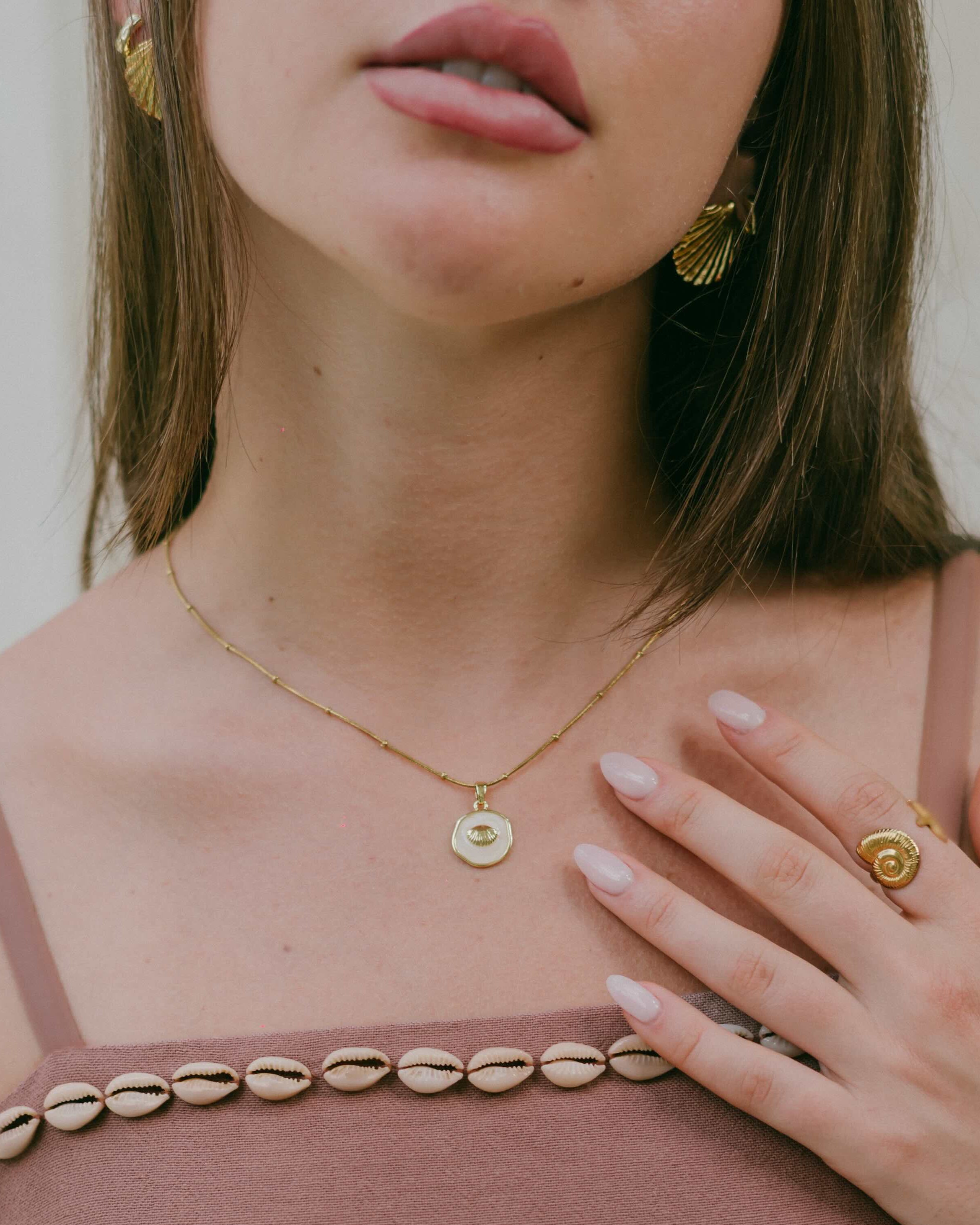 Shop the Naia Clam Necklace—an 18k gold plated stainless steel waterproof necklace featuring a glossy shell-look circular pendant framed by a clam shell design. Perfect for layering or everyday wear.