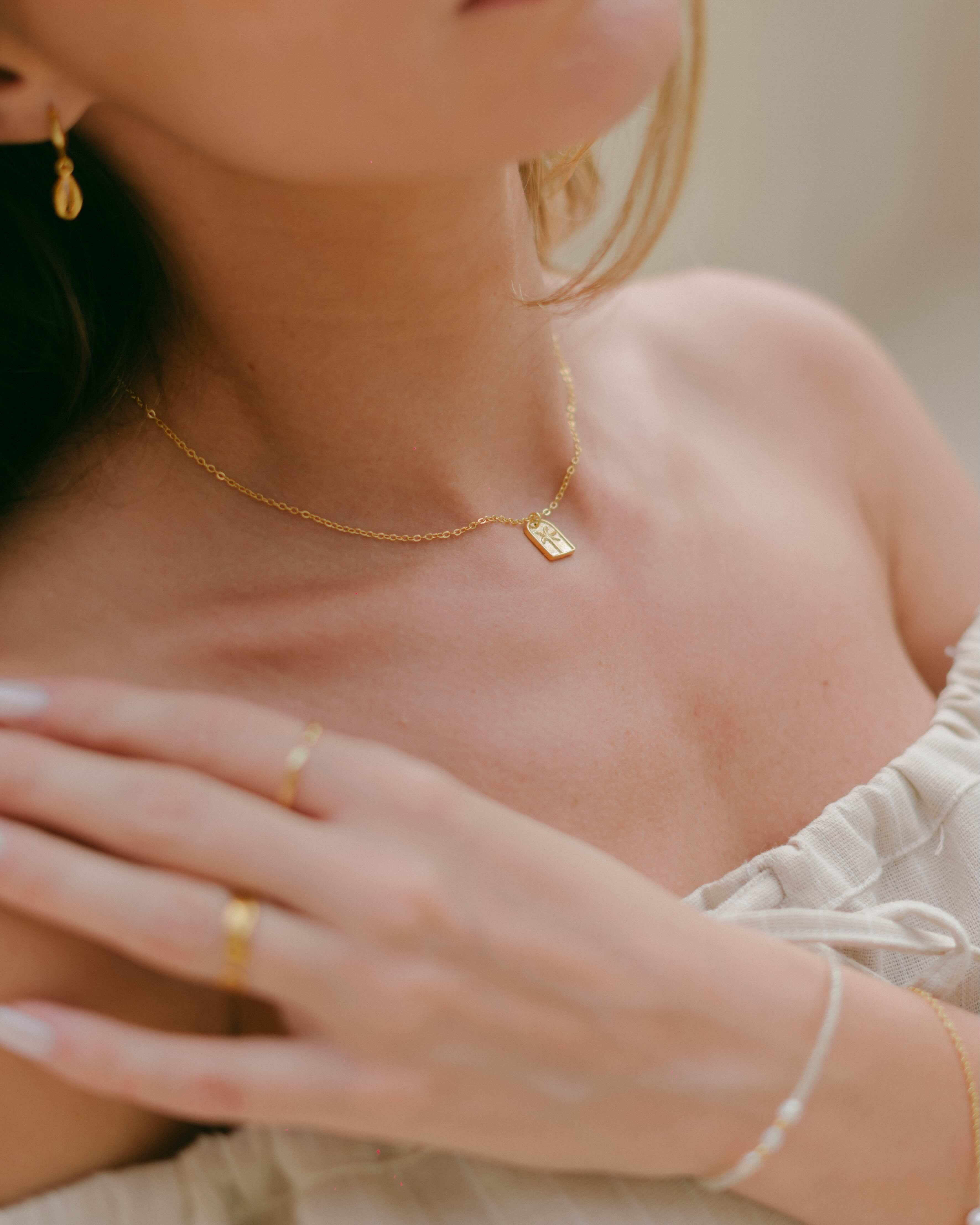 Elevate your look with the Isola Gold Necklace, showcasing a minimalist arched pendant with a serene palm tree engraving. A timeless, versatile gold necklace ideal for tropical-inspired fashion.