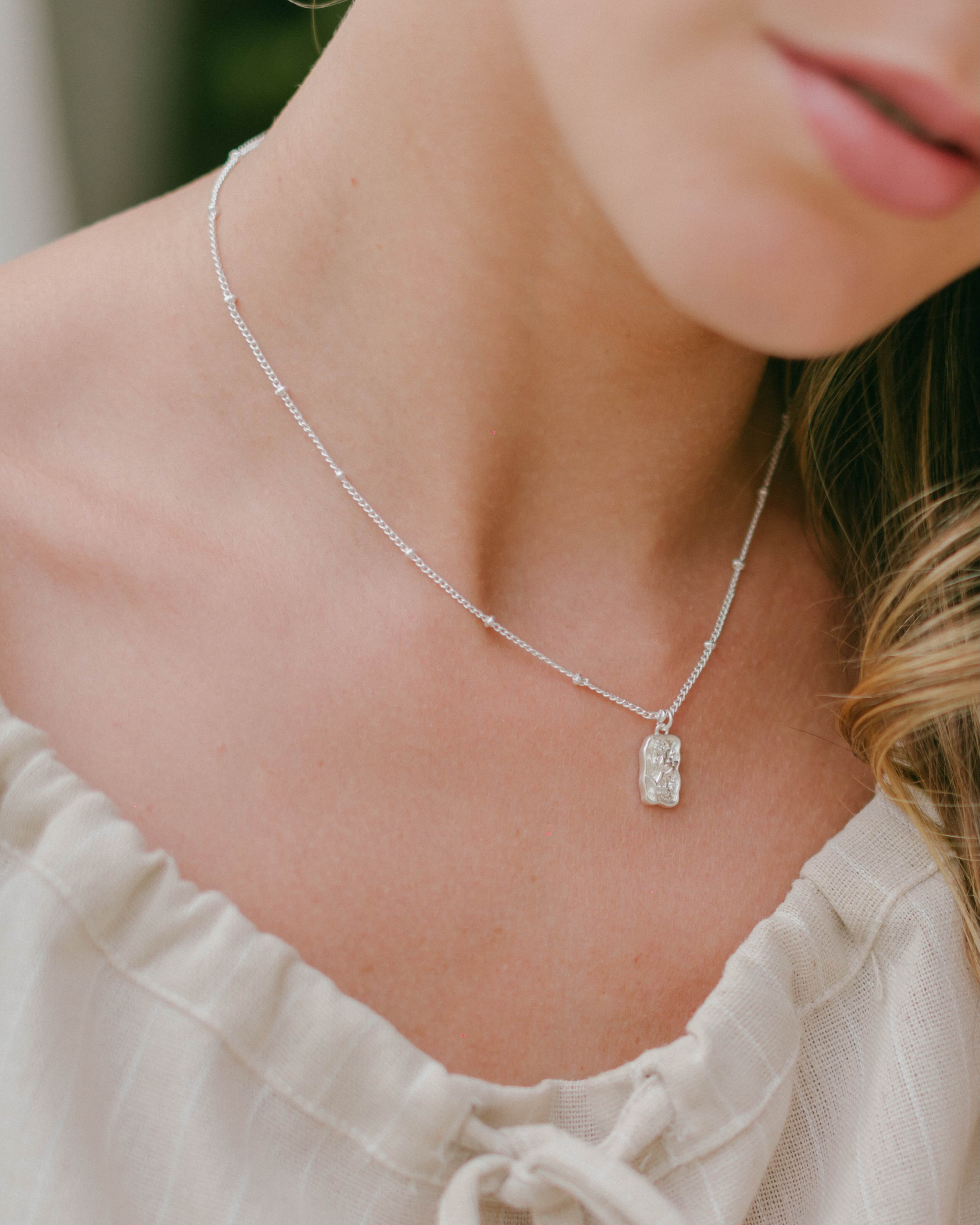 Shop the Flora Rectangle Necklace— a silver chain with detailed circular links and a small rectangular flower pendant. A delicate, versatile piece perfect for everyday wear and layering.
