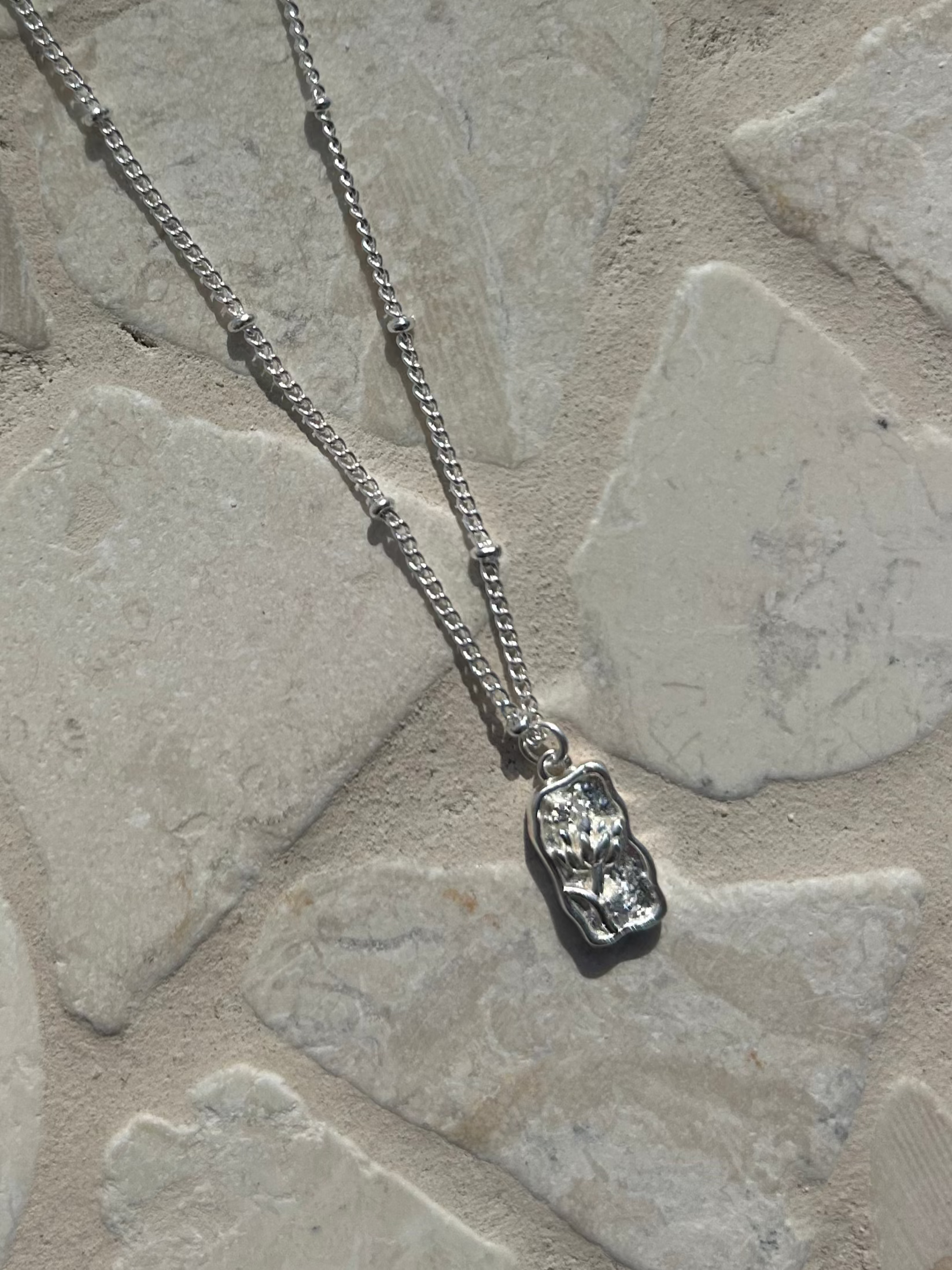 Discover the perfect blend of texture and floral charm with the Flora Rectangle Necklace. Silver chain with detailed circles and a flower-engraved pendant makes it a timeless accessory for any look.