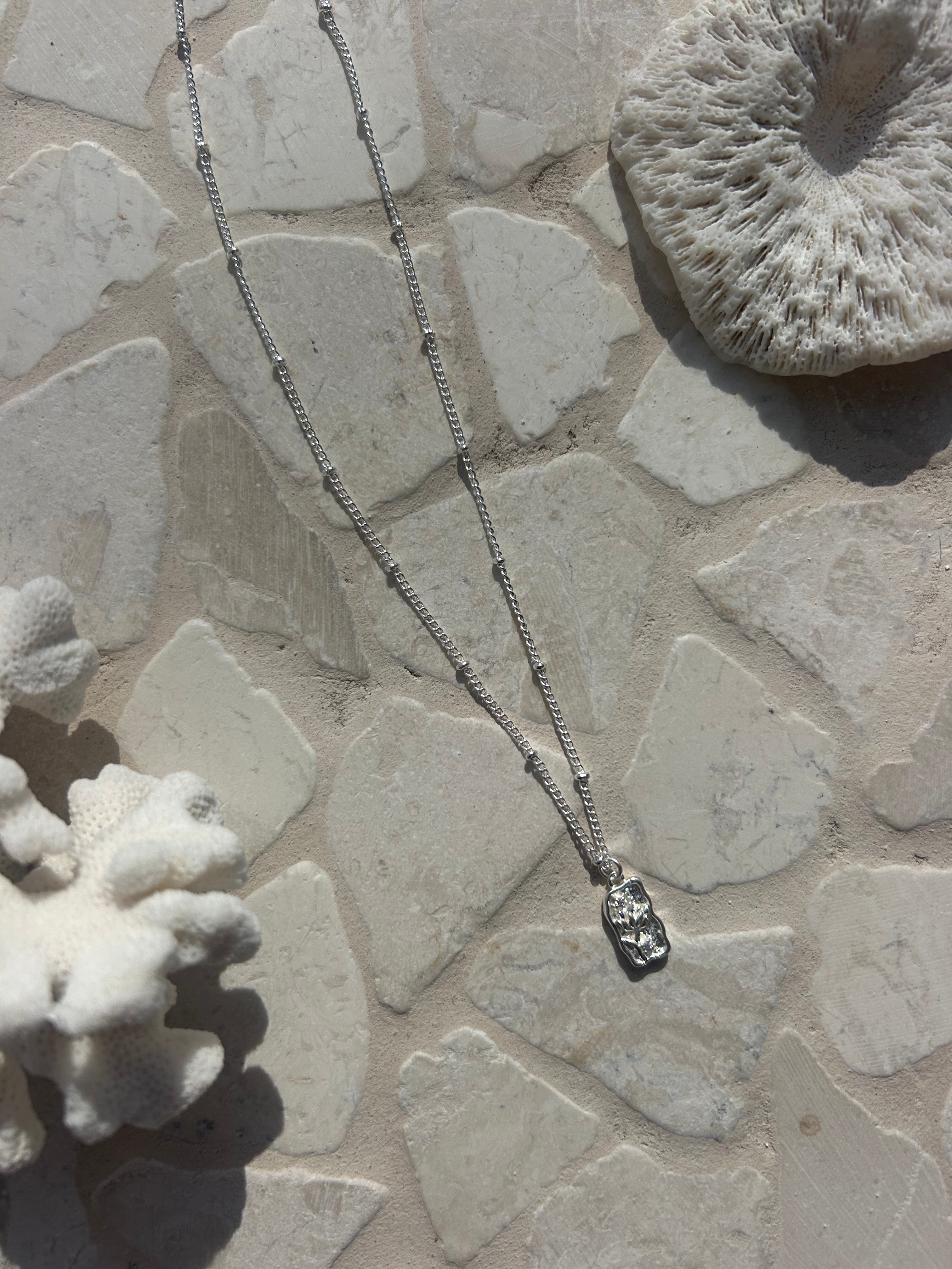 Elevate your jewellery collection with the Flora Necklace, featuring a silver chain decorated with intricate circle details and a floral rectangular pendant. Ideal for subtle elegance and daily styling.