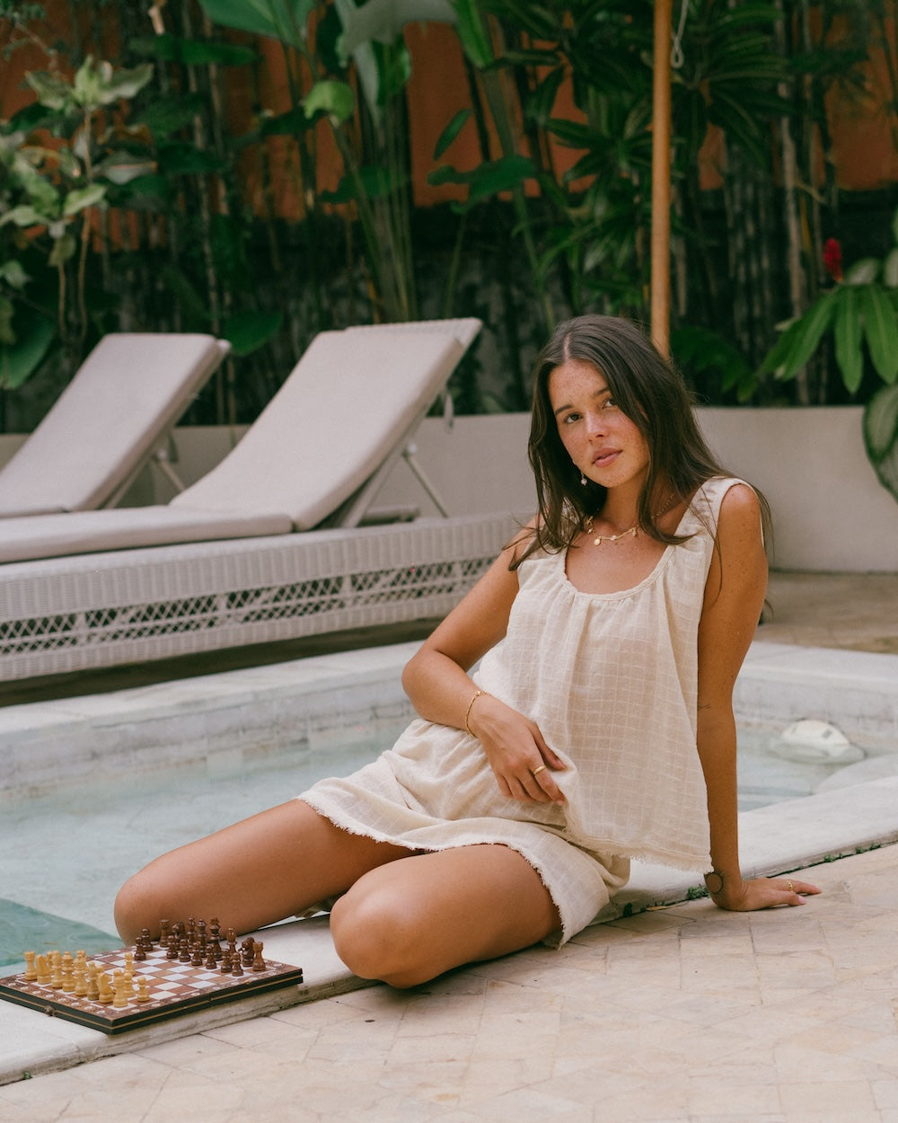 Sofia Singlet Top and Mini Skirt lifestyle shot — model seated on pool edge with chess board, full co-ord matching set visible, ruffled neckline and frayed hem detail — natural ramie cotton summer co-ord — Malia The Label La Dolce Vita