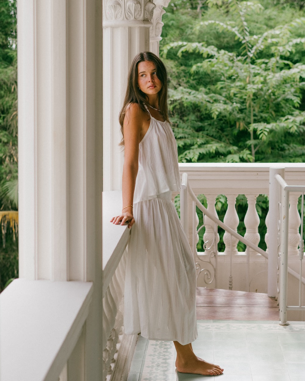Sole Mare Singlet Top and Maxi Skirt side profile on villa balcony — model leaning against white pillar, floor-length white co-ord set showing gathered halter top and maxi skirt drape, lush green backdrop — natural cotton slow fashion co-ord — Malia The Label La Dolce Vita