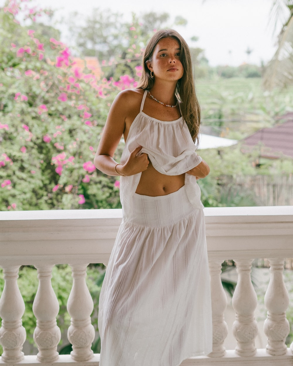 Sole Mare Singlet Top and Maxi Skirt lifestyle shot on balcony — model lifting singlet hem to show waist, full matching white set visible with gathered halter top and maxi skirt, pink bougainvillea and tropical backdrop — 100% cotton slow fashion co-ord — Malia The Label La Dolce Vita