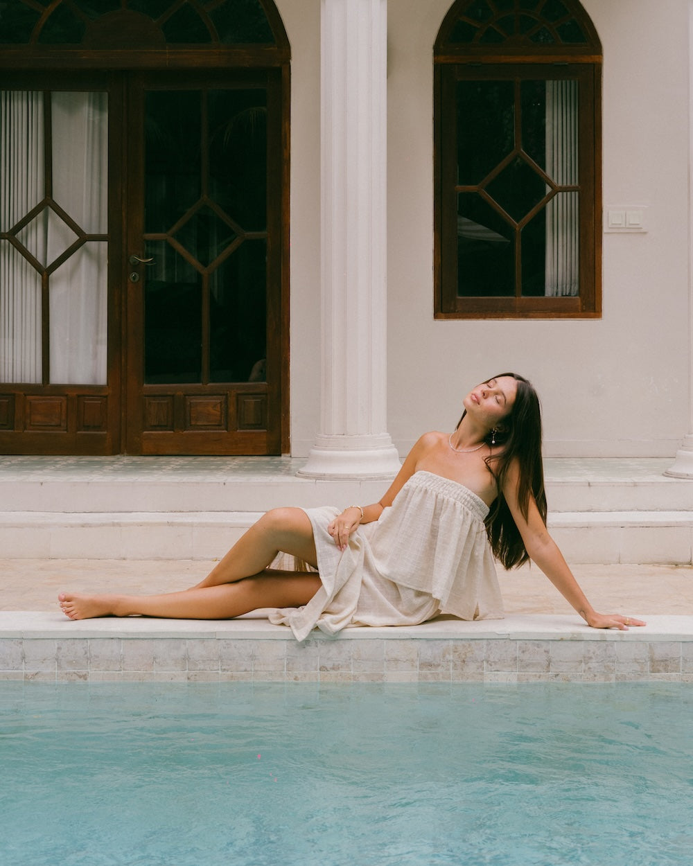 Sorrento Strapless Top worn solo poolside — model reclined on pool edge with eyes closed in warm light, shirred bandeau top with flowy gathered silhouette, white villa backdrop — natural ramie strapless top Australia — Malia The Label La Dolce Vita collection