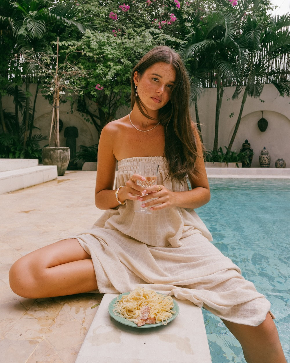 Sorrento Strapless Top and Maxi Skirt styled by pool — seated portrait showing shirred bandeau top and maxi skirt in textured check ramie, model looking directly at camera, pool tile background — natural ramie co-ord matching set — Malia The Label La Dolce Vita