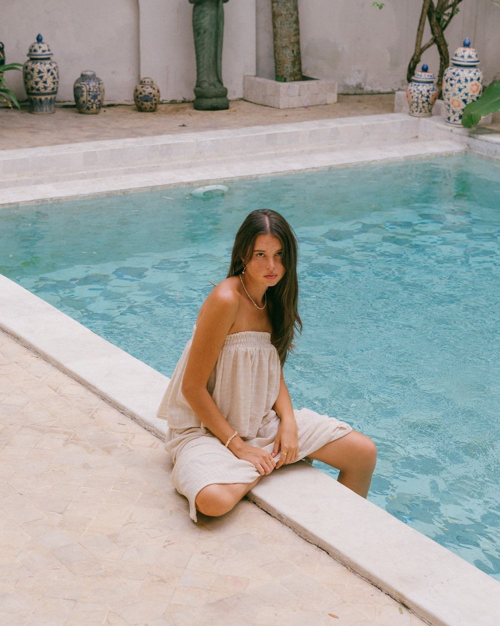 Sorrento Strapless Top and Maxi Skirt natural ramie co-ord — model kneeling at pool edge in full matching set, strapless shirred bandeau and floor-length skirt, blue and white ceramic vases in background — natural fibre matching set — Malia The Label La Dolce Vita