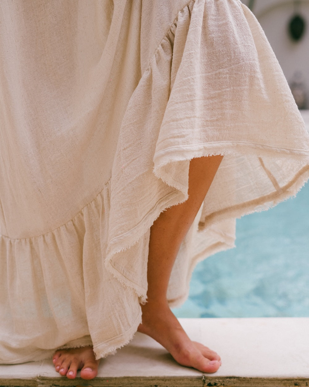 Vela Maxi Dress ruffled hem close-up detail — frayed natural ramie fabric caught mid-movement poolside, barefoot model, showing textured weave and tiered ruffle — 80% ramie 20% cotton maxi dress hem detail — Malia The Label slow fashion