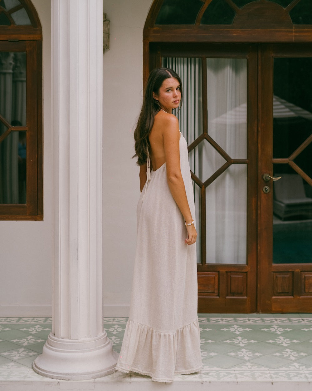 Vela Maxi Dress backless halter style — model looking over shoulder, three-quarter rear view showing open back, braided tie at neckline and full-length ruffled hem — natural ramie cotton backless maxi dress — Malia The Label Australia