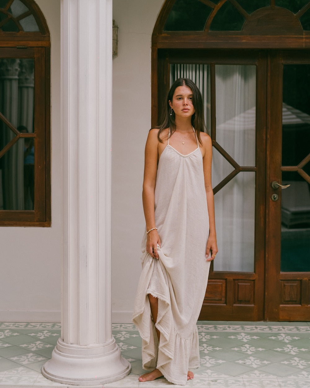 Vela Maxi Dress front full-length view — model standing by white column and timber door, braided v-neckline and free-flowing silhouette clearly visible, ruffled hem grazing tiled floor — natural ramie cotton maxi dress — Malia The Label