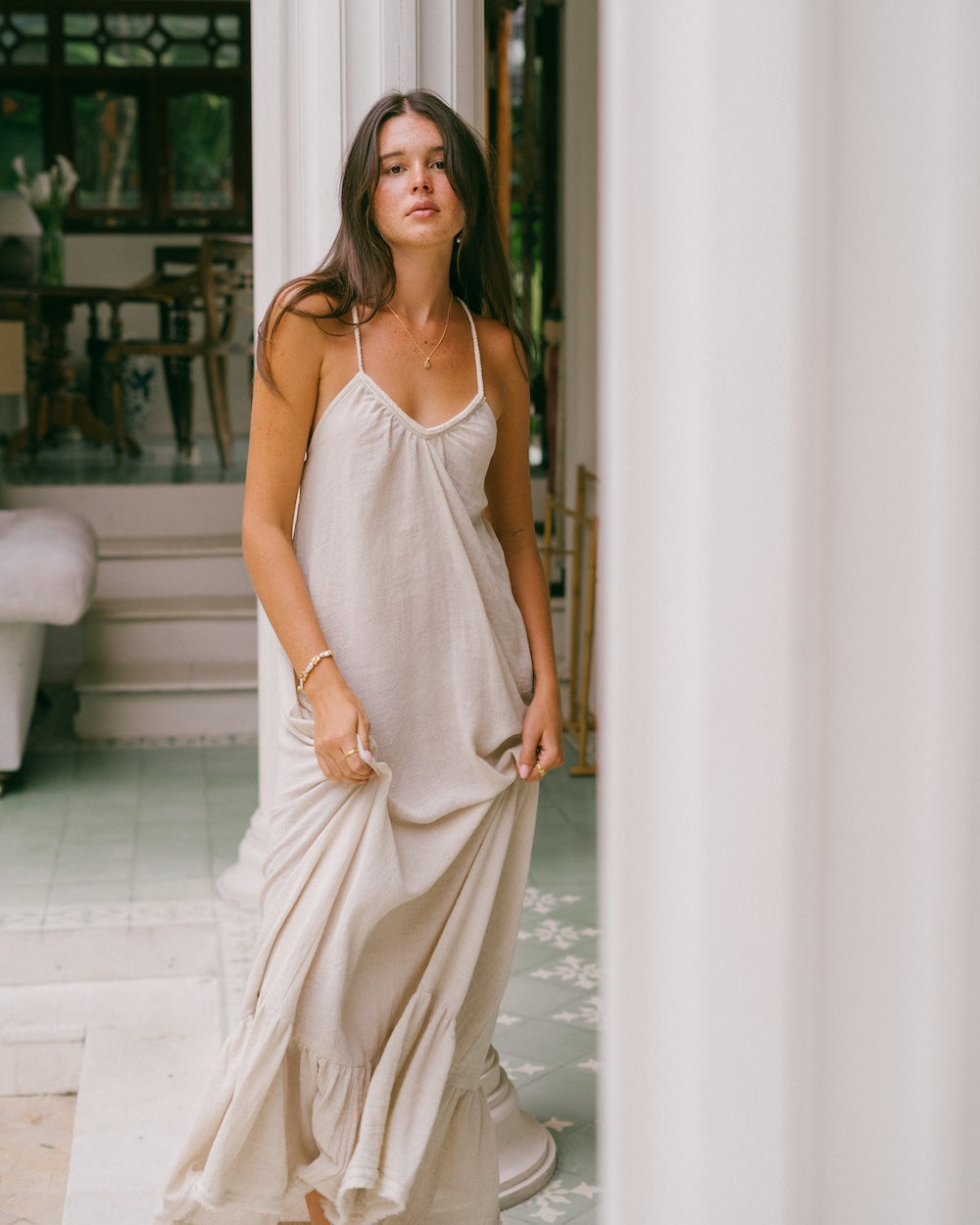 Vela Maxi Dress in natural ramie — model standing between white columns lifting fabric to show free-flowing drape and movement, braided v-neckline detail visible — handloomed natural fibre maxi dress — Malia The Label La Dolce Vita collection