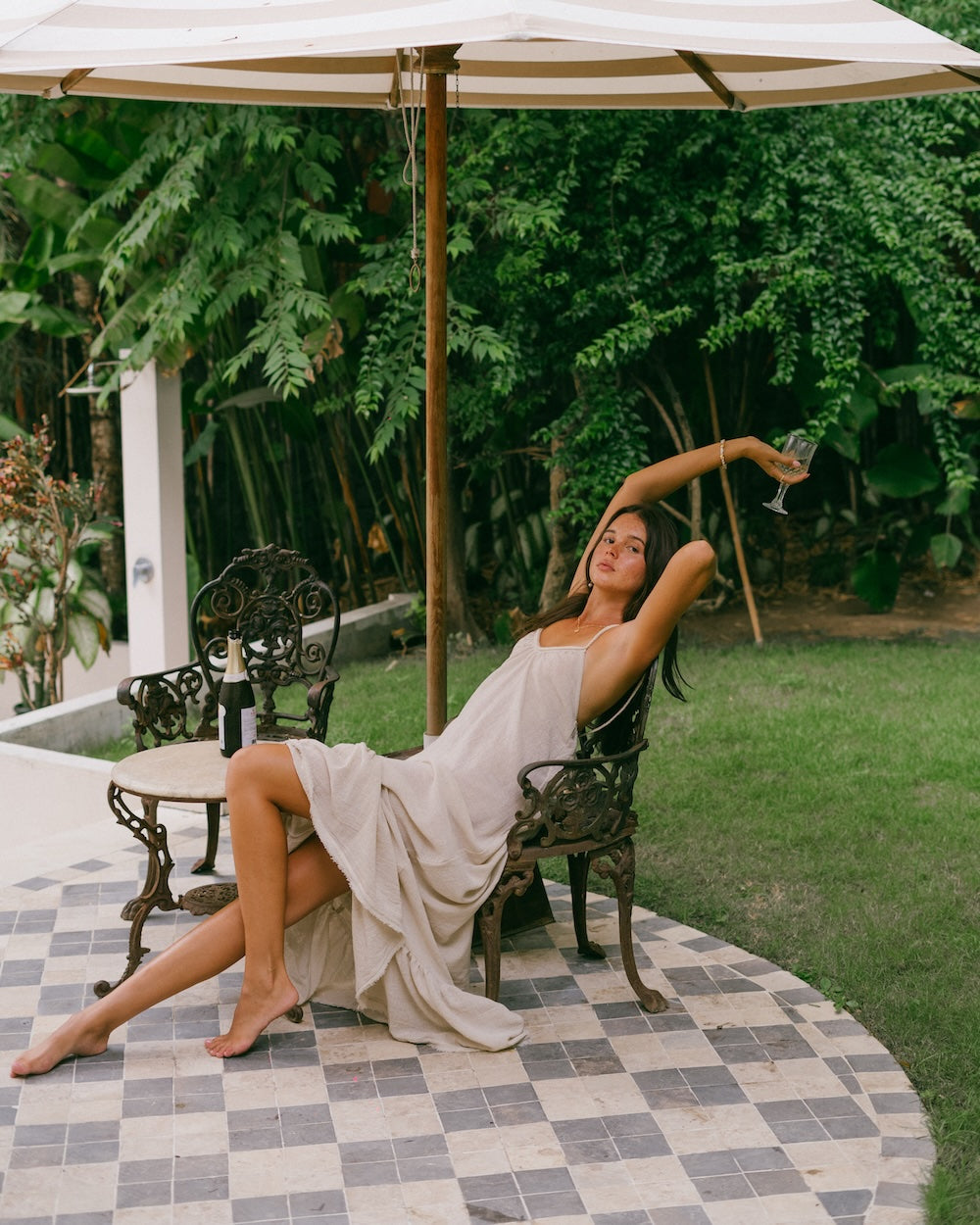 Vela Maxi Dress lifestyle shot — model relaxed in ornate cast iron garden chair under striped umbrella holding a wine glass, natural ramie dress draped effortlessly — slow living summer style — Malia The Label La Dolce Vita collection Australia