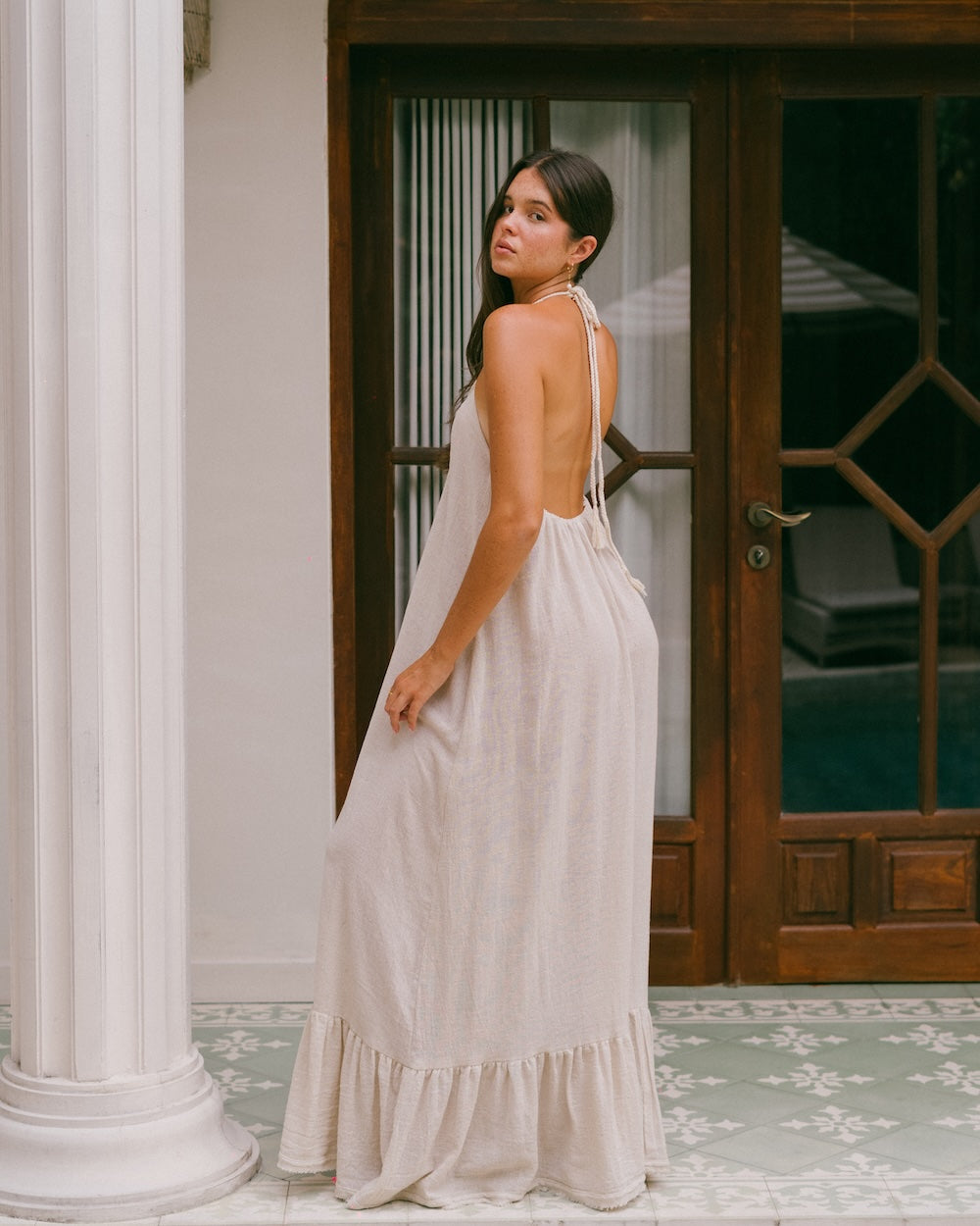 Vela Maxi Dress side halter back variation — three-quarter side view showing braided halter strap tied at nape of neck, open back and free-flowing natural ramie skirt, ruffled hem grazing tiled floor — convertible backless halter maxi dress — Malia The Label La Dolce Vita collection — natural fibre slow fashion Australia