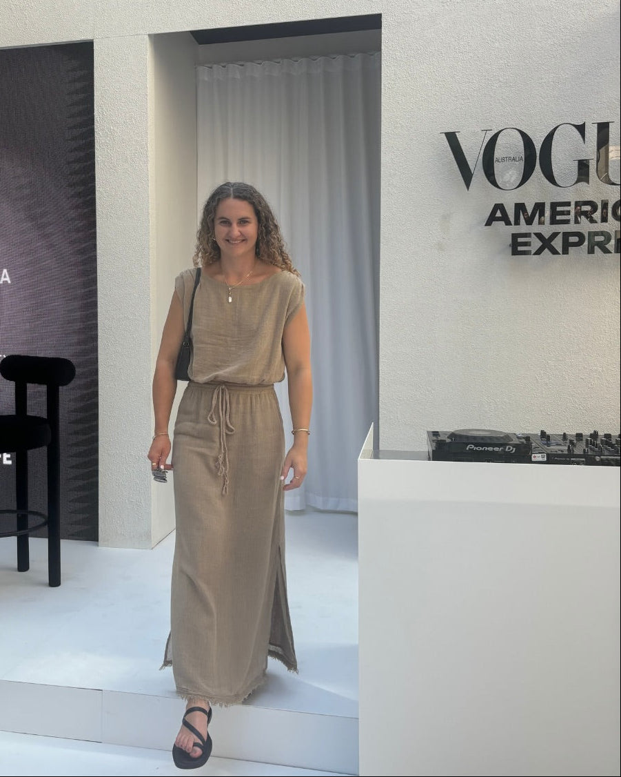 A woman stands on a white stage at a Vogue America Express event, wearing a beige sleeveless maxi dress with a drawstring waist. The background features a white wall with the text 'What if I never say yes to taking the risk?' and 'VOGUE AMERICA EXPRESS', alongside a black chair and a dark panel with 'BELOW THE HAT' and 'SEPHORA' branding.